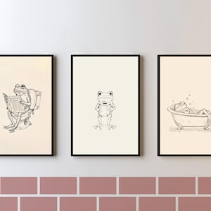 Può includere: Un set di tre stampe da parete incorniciate con disegni a linee nere di rane su sfondo color crema. La prima mostra una rana sul water che legge un giornale, la seconda una rana che tiene un rotolo di carta igienica e la terza una rana in una vasca da bagno.
