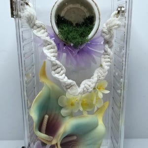 May include: A clear plastic terrarium with decorative elements. It contains a white rope, artificial flowers, and calla lilies in yellow, purple, and green. A small pot with greenery and pink flowers is at the top.
