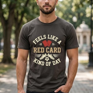 May include: A dark gray short-sleeved t-shirt featuring a graphic print with the text "FEELS LIKE A RED CARD KIND OF DAY" in beige, two red rectangular cards, a soccer ball, and two white soccer cleats arranged in a centered design.