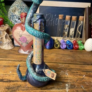 Pode incluir: Uma garrafa de poção azul escuro com uma cobra azul e turquesa enrolada ao redor. A garrafa tem um rótulo que diz "Veneno de Cobra". Uma pequena etiqueta pende da garrafa. A garrafa está em uma superfície de madeira.