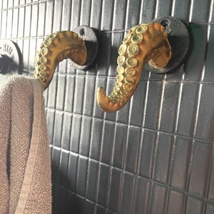 Può includere: Due ganci a forma di tentacolo di polpo color bronzo montati su una parete piastrellata grigio scuro. Un gancio tiene un asciugamano beige. I ganci hanno un design dettagliato e strutturato e sono attaccati a basi circolari grigio scuro.