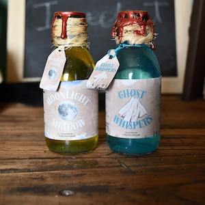 Puede incluir: Dos botellas decorativas de vidrio con etiquetas que dicen "Moonlight Shadow" y "Ghost Whispers". Las botellas tienen sellos de cera y etiquetas colgantes. La botella "Moonlight Shadow" contiene un líquido amarillo, mientras que la botella "Ghost Whispers" contiene un líquido azul.