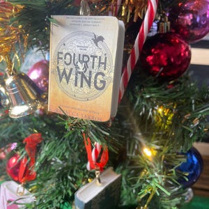 Può includere: Un albero di Natale decorato con ornamenti, tra cui un libro in miniatura intitolato "Fourth Wing" di Rebecca Yarros. Il libro è beige con scritte nere e un design circolare. Altri ornamenti includono un bastoncino di zucchero, campane e palline colorate.