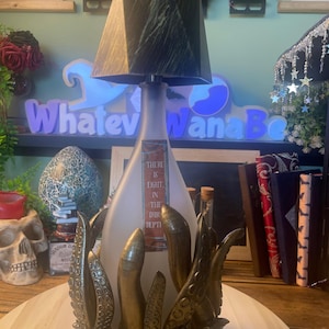 Può includere: Una lampada unica con un paralume geometrico nero e una base in vetro smerigliato. La base è adornata da tentacoli color bronzo. La lampada presenta il testo "THERE IS LIGHT IN THE DARK DEPTHS".