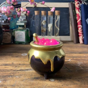 Può includere: Un piccolo calderone viola scuro con bordo dorato e dettagli dorati gocciolanti. Il calderone è pieno di forme rosa brillante a forma di bolla e ha un piccolo cucchiaio d'oro. Il calderone è su una superficie di legno.
