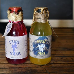 Könnte beinhalten: Zwei dekorative Glasflaschen mit farbigen Flüssigkeiten. Eine Flasche ist mit einer roten Flüssigkeit gefüllt und mit "Fairy Sugar" beschriftet. Die andere Flasche enthält eine gelbe Flüssigkeit und ist mit "Pixie Dust" beschriftet. Beide Flaschen haben dekorative Etiketten und sind mit Wachs versiegelt.