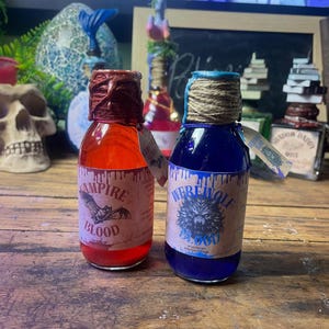 May include: Two glass bottles with labels reading "Vampire Blood" and "Werewolf Blood." The red liquid bottle has a brown wax seal, while the blue liquid bottle has a twine-wrapped neck. Both bottles have decorative tags.