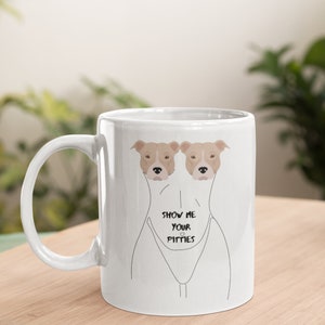 Pode incluir: Caneca de cerâmica branca com um contorno preto de uma pessoa com duas cabeças de pit bull espiando do peito. O texto "Show me your pitties" está impresso na caneca.