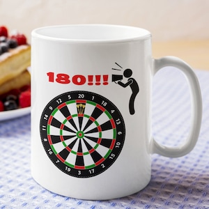 May include: White ceramic mug featuring a dartboard design with the number "180!!!" in red. A stick figure is depicted throwing darts. The mug has a curved handle and is suitable for hot or cold beverages.