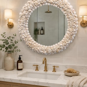 May include: A round mirror with a frame made of seashells hangs above a bathroom sink. The sink has a gold faucet and fixtures. A vase with greenery and a soap dispenser are on the countertop. Two wall sconces flank the mirror.