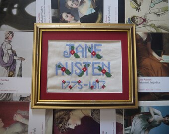 Op de afbeelding: Een ingelijst borduurwerk met kruissteek met de tekst "JANE AUSTEN 1775-1817" in blauwe letters met rode bloemen en groene bladeren op een witte achtergrond. De lijst is goudkleurig met een rode passe-partout.