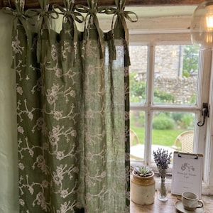May include: An olive green curtain panel featuring a delicate white floral embroidery pattern. The curtain is suspended from a dark wooden rod using five fabric tie-top attachments. The fabric is semi-sheer, allowing light to pass through the floral design.