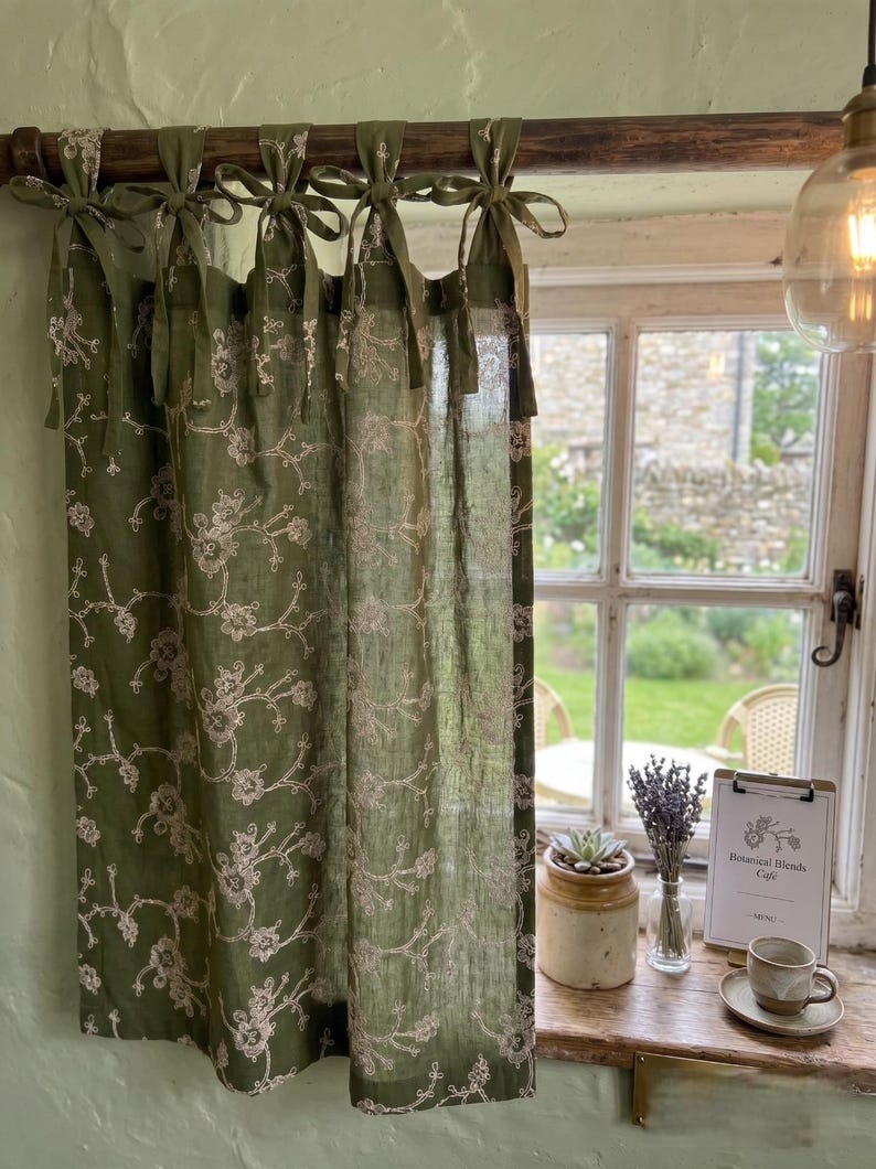 May include: An olive green curtain panel featuring a delicate white floral embroidery pattern. The curtain is suspended from a dark wooden rod using five fabric tie-top attachments. The fabric is semi-sheer, allowing light to pass through the floral design.