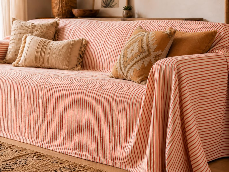 K&ouml;nnte beinhalten: Ein Sofa, das mit einer Stoffdecke mit d&uuml;nnen vertikalen orangefarbenen und wei&szlig;en Streifen bedeckt ist. Das Sofa ist mit vier dekorativen Kissen in verschiedenen Braun- und Beiget&ouml;nen geschm&uuml;ckt, darunter eines mit geometrischem Muster und Fransen.