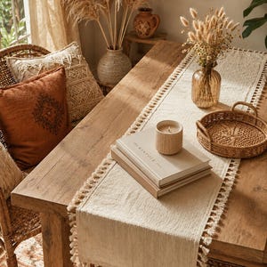 May include: A rectangular off-white fabric table runner featuring a textured weave and decorative fringe trim along both long edges. The runner is placed on a wooden table, topped with two stacked books, a candle, and a small woven basket.