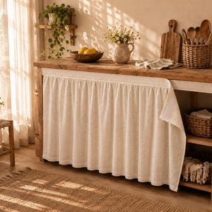 May include: A wooden kitchen island featuring a beige fabric skirt attached with hook-and-loop fasteners. The island has an open shelving unit on the right side containing two woven baskets, while the top surface holds a bowl of lemons and a vase.