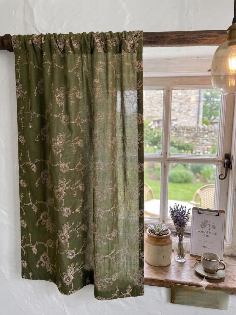 May include: An olive green curtain featuring a delicate, light-colored floral embroidery pattern. The fabric is semi-sheer and hangs from a dark wooden rod, displaying a repeating vine and flower motif across the entire surface of the panel.