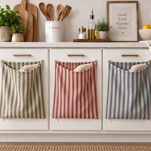 May include: Three rectangular fabric storage pouches with vertical stripes in olive green, red, and blue, each suspended from gold-toned hardware on white kitchen cabinet doors. Each pouch contains a small, light-coloured textured cloth item.