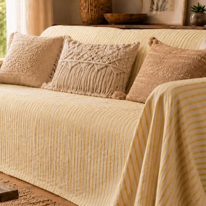 K&ouml;nnte beinhalten: Ein Sofa mit einem hellgelb-wei&szlig; gestreiften Bezug, dekoriert mit drei Zierkissen. Ein Kissen hat ein strukturiertes geometrisches Muster, ein weiteres ist ein cremefarbenes Makramee-Design mit Fransen, und das dritte ist ein beiges Webkissen.