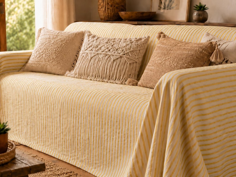 K&ouml;nnte beinhalten: Ein Sofa mit einem hellgelb-wei&szlig; gestreiften Bezug, dekoriert mit drei Zierkissen. Ein Kissen hat ein strukturiertes geometrisches Muster, ein weiteres ist ein cremefarbenes Makramee-Design mit Fransen, und das dritte ist ein beiges Webkissen.