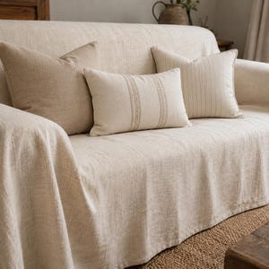 Könnte beinhalten: Ein beiges Sofa, das mit einer strukturierten, hellen Tagesdecke bedeckt ist. Das Sofa ist mit drei Zierkissen ausgestattet: einem einfarbigen beigen quadratischen Kissen, einem rechteckigen Kissen mit einem mittigen vertikalen Stickstreifen und einem quadratischen Kissen mit feinen vertikalen Streifen.