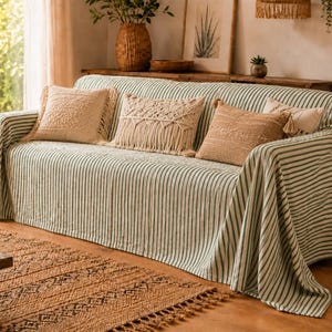 Puede incluir: Un sofá cubierto con una manta de tela a rayas de color verde claro y blanco. El sofá está adornado con tres cojines decorativos, incluidos dos cojines beige texturizados y un cojín rectangular central con detalles de flecos de macramé.