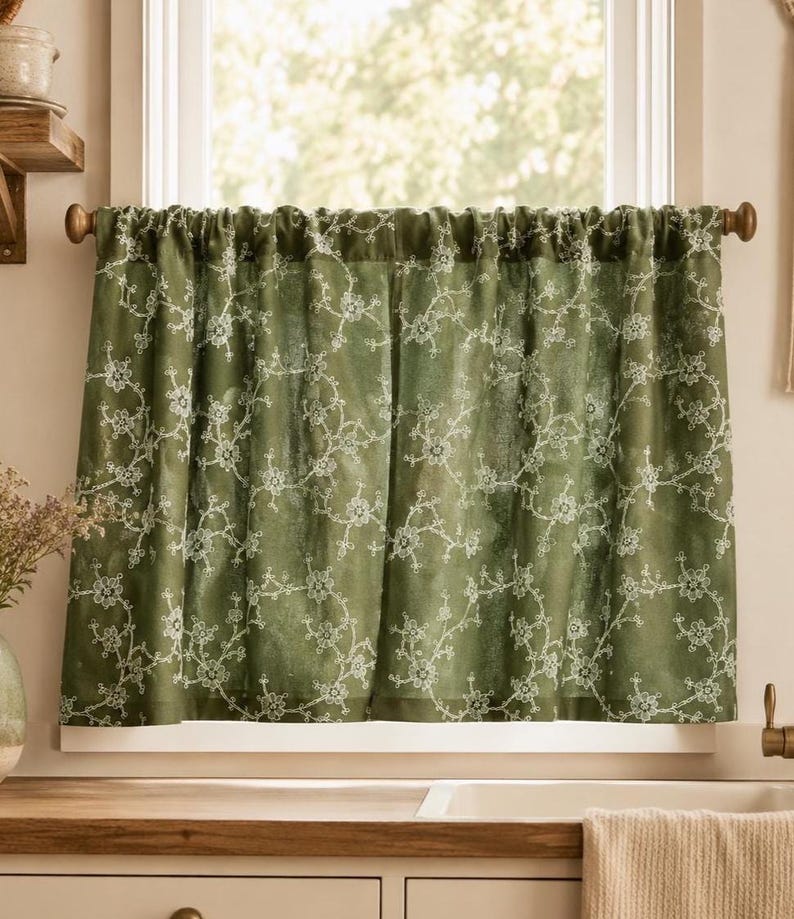 May include: A sage green cafe curtain featuring a delicate white embroidered floral vine pattern. The curtain is gathered on a wooden rod and hangs over a window, displaying a semi-sheer fabric with intricate stitched details throughout the design.