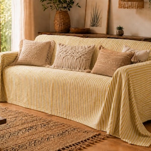K&ouml;nnte beinhalten: Ein Sofa, das mit einer gelb-wei&szlig; gestreiften Stoffdecke bedeckt ist und drei dekorative Kissen aufweist. Zwei Kissen sind beige mit strukturierten Mustern, und das mittlere Kissen ist cremefarben mit Makramee-Fransen.