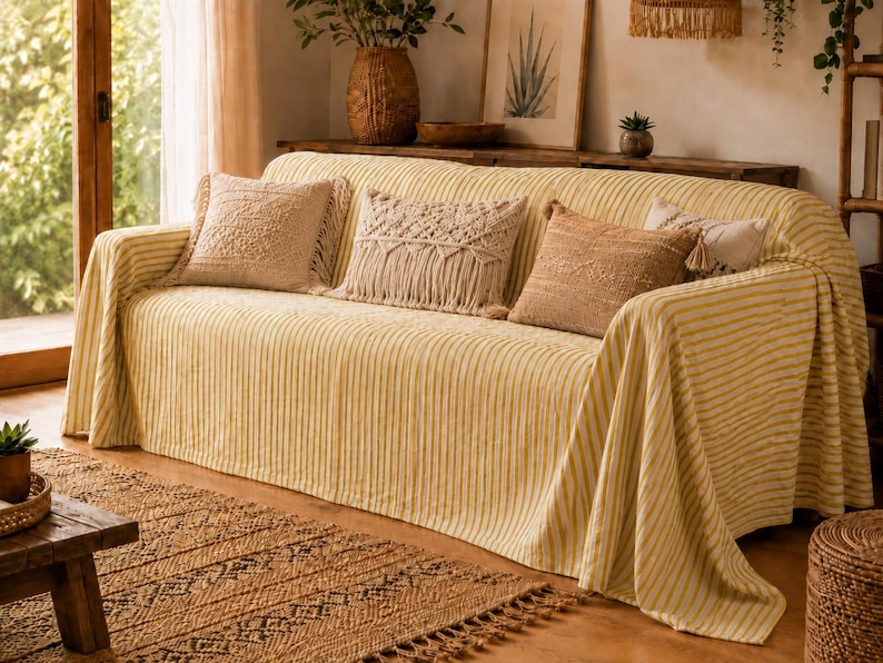 K&ouml;nnte beinhalten: Ein Sofa, das mit einer gelb-wei&szlig; gestreiften Stoffdecke bedeckt ist und drei dekorative Kissen aufweist. Zwei Kissen sind beige mit strukturierten Mustern, und das mittlere Kissen ist cremefarben mit Makramee-Fransen.