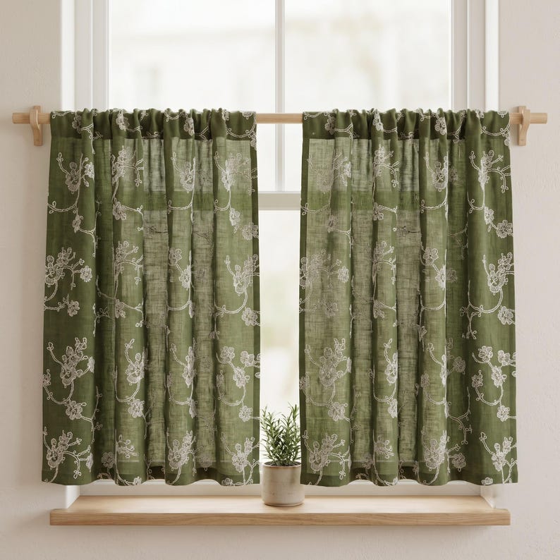May include: A pair of olive green cafe curtains featuring a white embroidered floral vine pattern. The curtains are hung on a wooden rod and have a rod pocket header, displaying a semi-sheer fabric texture that allows light to pass through.