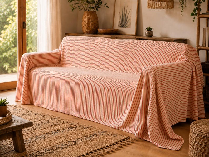 K&ouml;nnte beinhalten: Ein Sofa, das mit einer hellpfirsichfarbenen und wei&szlig;en gestreiften Stoffdecke bedeckt ist. Das Textil weist eine subtile vertikale Rippenstruktur auf und f&auml;llt &uuml;ber die Armlehnen und die R&uuml;ckenlehne des M&ouml;bels, was eine weiche Optik erzeugt.