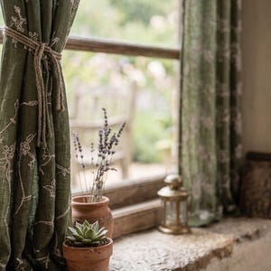 May include: Olive green curtains featuring a delicate white floral embroidery pattern, held back by a simple jute rope tie. The fabric is semi-sheer and hangs in soft folds, framing a window view that includes a small succulent in a terracotta pot.