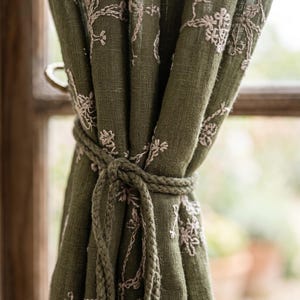 May include: An olive green curtain panel featuring a delicate, light-colored floral embroidery pattern. The fabric has a visible, textured weave and is gathered at the center by a matching braided olive green rope tieback, creating soft folds.