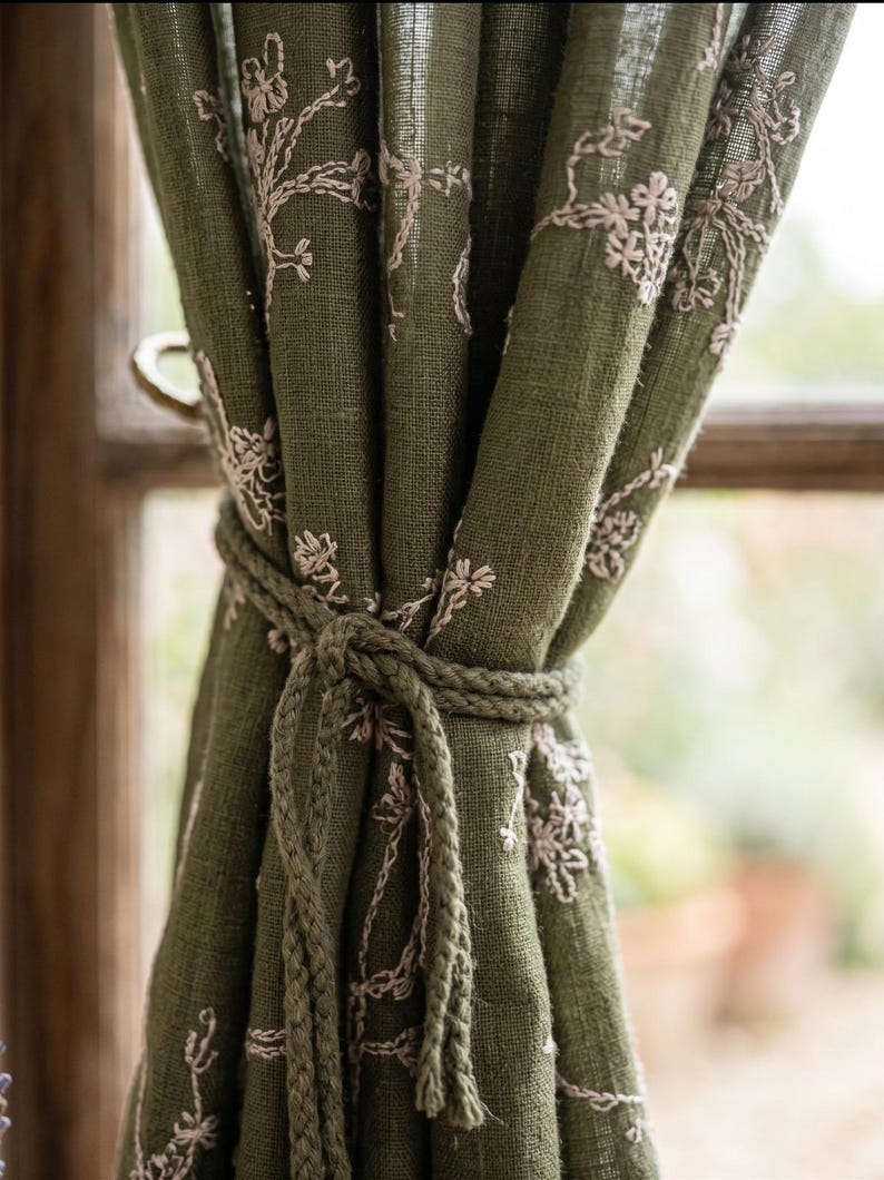 May include: An olive green curtain panel featuring a delicate, light-colored floral embroidery pattern. The fabric has a visible, textured weave and is gathered at the center by a matching braided olive green rope tieback, creating soft folds.