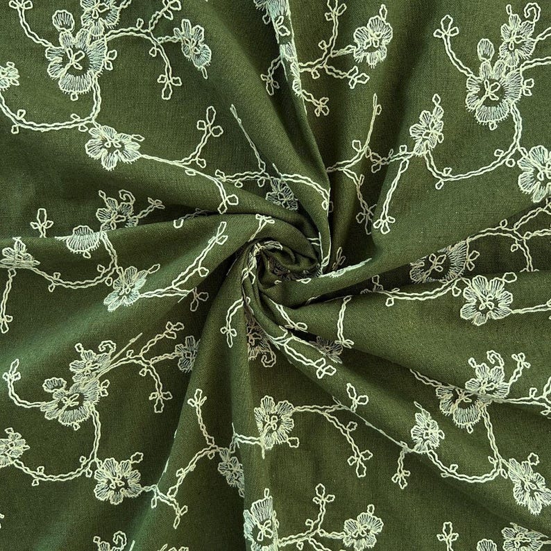 May include: An olive green fabric featuring a delicate white embroidered floral pattern. The design consists of thin, winding branches with small blossoms and leaves scattered across the surface, creating a continuous, intricate botanical motif.
