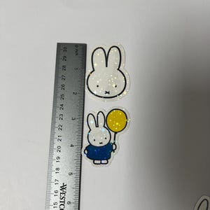 May include: Three holographic stickers featuring a white rabbit with black outlines. The first sticker shows the rabbit's head with a white background. The second sticker shows the rabbit holding a yellow balloon with a blue shirt and white background. The third sticker shows the rabbit with an orange shirt and white background.