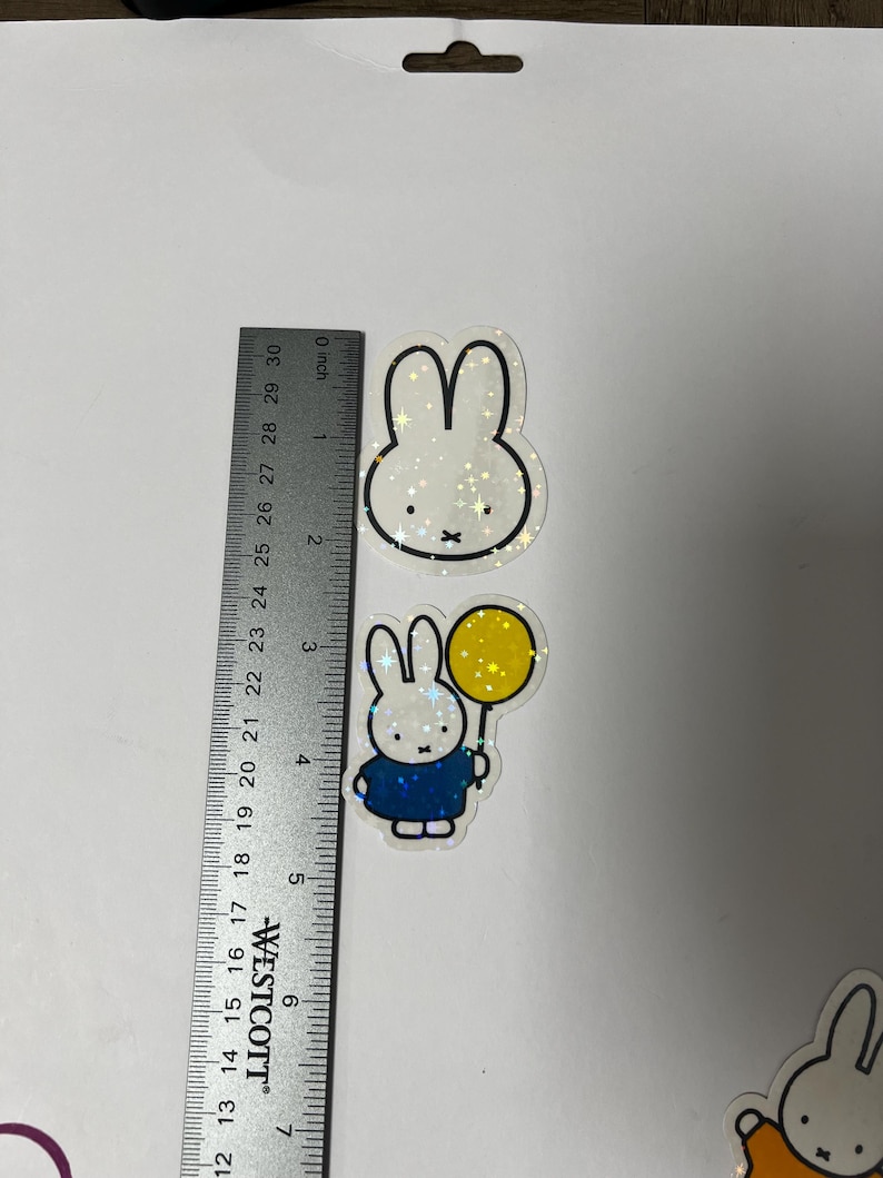 May include: Three holographic stickers featuring a white rabbit with black outlines. The first sticker shows the rabbit's head with a white background. The second sticker shows the rabbit holding a yellow balloon with a blue shirt and white background. The third sticker shows the rabbit with an orange shirt and white background.