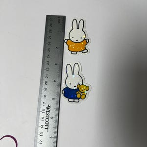 May include: Two white bunny stickers with glitter accents. The first bunny is wearing a yellow shirt. The second bunny is wearing a blue shirt and holding a yellow teddy bear.