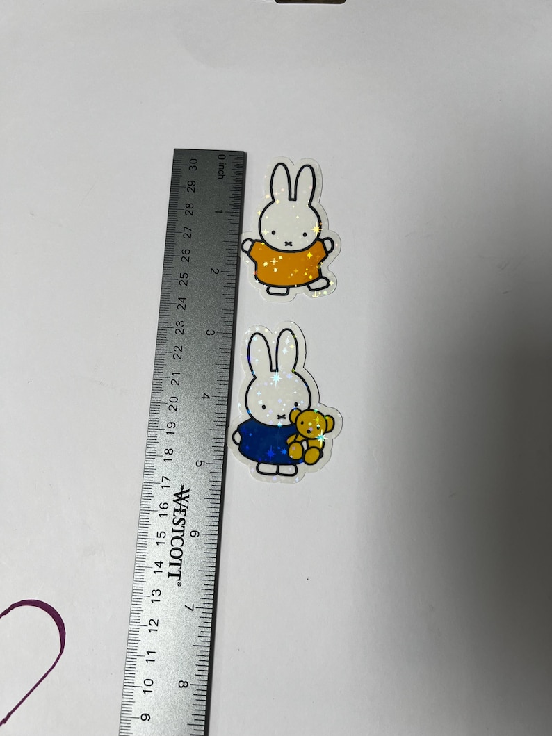 May include: Two white bunny stickers with glitter accents. The first bunny is wearing a yellow shirt. The second bunny is wearing a blue shirt and holding a yellow teddy bear.