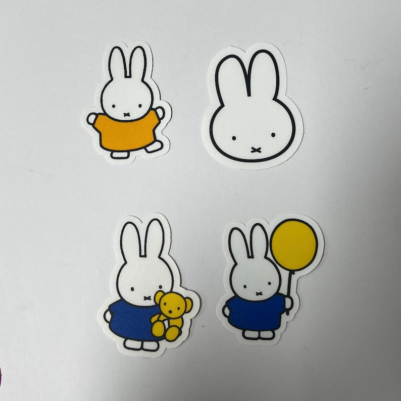 May include: Four white bunny stickers with black outlines. One bunny is wearing a yellow shirt, one is holding a yellow balloon, one is holding a yellow teddy bear, and one is plain.