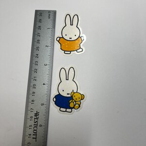 May include: Two white glitter bunny stickers with black outlines. The first bunny is wearing a yellow shirt and the second bunny is wearing a blue shirt and holding a yellow teddy bear.