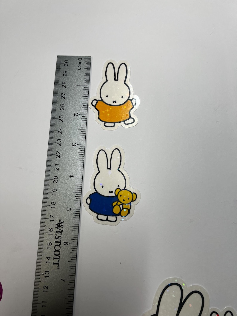 May include: Two white glitter bunny stickers with black outlines. The first bunny is wearing a yellow shirt and the second bunny is wearing a blue shirt and holding a yellow teddy bear.