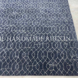 May include: A rectangular, dark blue rug with a repeating, interconnected, white geometric pattern. The rug has a textured surface and the text "THE HANDMADE RUGS IN" is printed across the bottom.