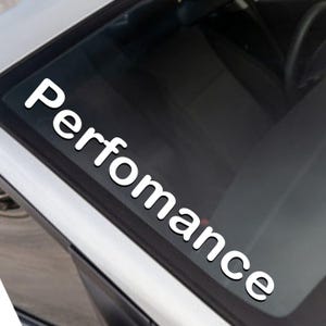 Può includere: Adesivo in vinile bianco con la parola "Performance" in un carattere audace e stilizzato. L'adesivo è applicato sul lunotto posteriore di un'auto.