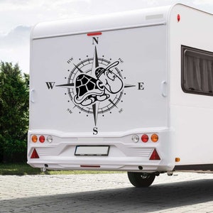 Könnte beinhalten: Ein weißer Wohnwagen mit einem schwarzen Kompass und einem Schildkröten-Design auf der Rückseite. Der Kompass zeigt nach Norden, Süden, Osten und Westen mit den Buchstaben N, S, O und W. Die Schildkröte blickt nach Osten und hat einen gepunkteten Panzer.