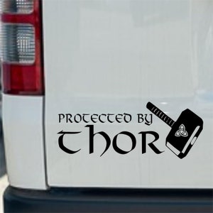 Op de afbeelding: Zwart-witte vinyl sticker met de tekst "Beschermd door Thor" in een gestileerd lettertype. Rechts van de tekst is een zwart silhouet van Thor's hamer, Mjolnir, afgebeeld. De hamer heeft een Keltisch knoopontwerp op zijn kop.