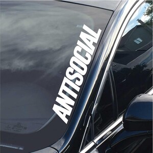 Pode incluir: Um adesivo de vinil branco com a palavra "ANTISOCIAL" escrita verticalmente no pára-brisas de um carro preto.
