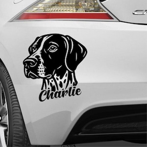 Könnte beinhalten: Ein schwarz-weißer Aufkleber mit dem Kopf eines Hundes und dem Namen "Charlie" in Schreibschrift darunter. Der Aufkleber befindet sich auf der weißen Karosserie eines Fahrzeugs.