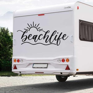 Op de afbeelding: Een witte camper met een zwarte vinyl sticker waarop "beachlife" staat met een zon en golven afbeelding. De camper heeft een kentekenplaat en achterlichten.