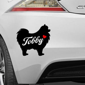 Op de afbeelding: Zwarte Pomeranian hondensticker met de naam "Tobby" in wit schrift en een klein rood hart. De sticker is bevestigd aan de achterkant van een wit voertuig.